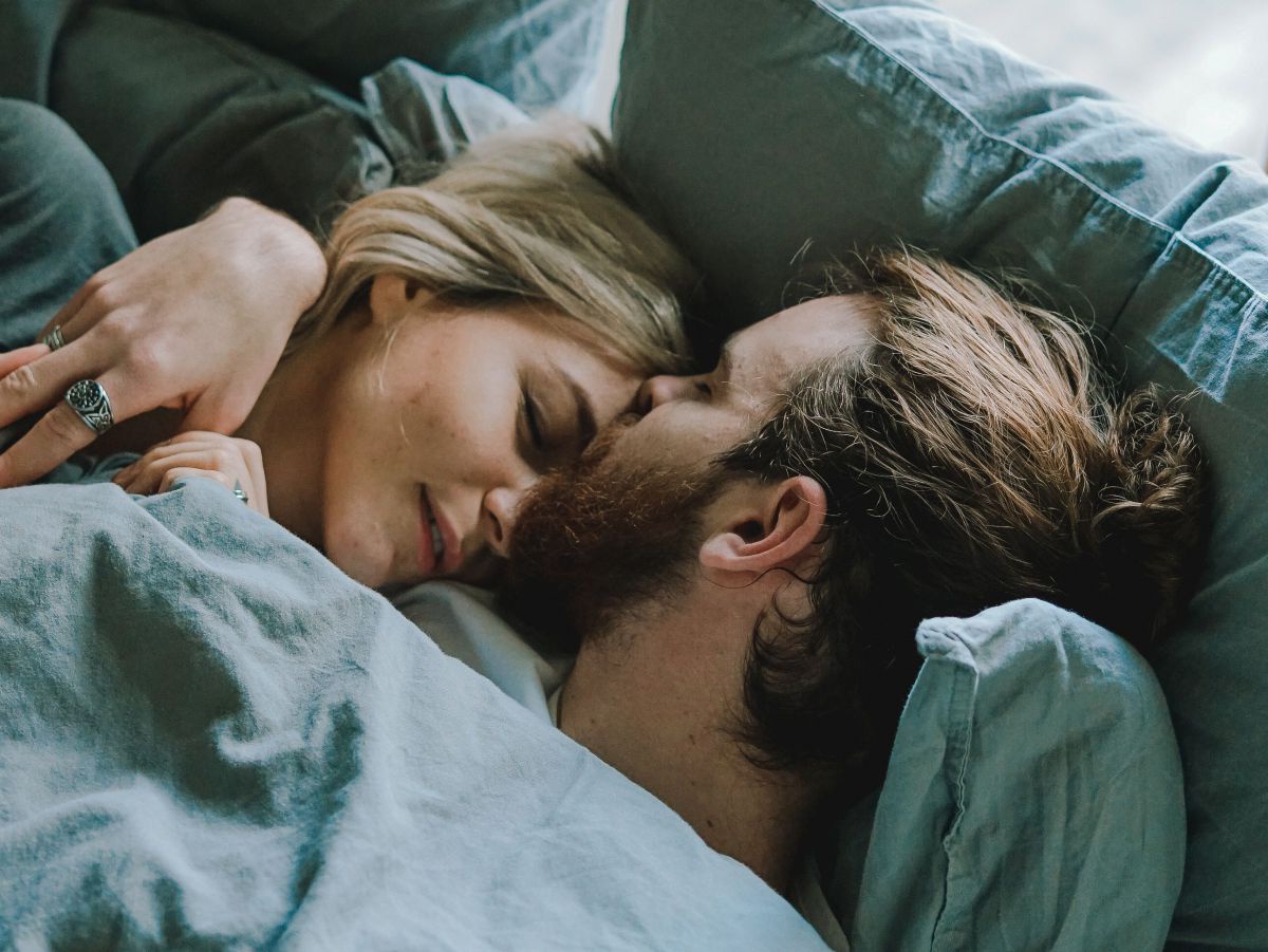 Ein heterosexuelles Paar kuschelnd in einem Bett in blauer Bettwäsche.