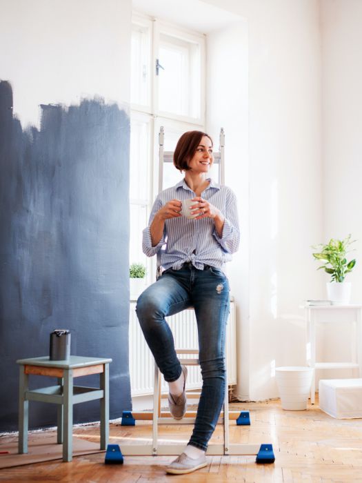 Eine Frau sitzt auf einer Haushaltsleiter in einem hohen Raum mit weißen Wänden. Sie macht gerade Pause, neben Ihr steht eine offene Farbdose auf einem Hocker und die Wand neben ihr ist zur Hälfte mit dunkler Farbe gestrichen.