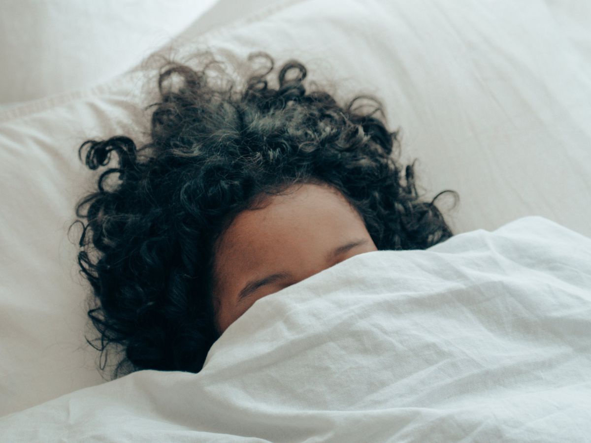 Ein Kind mit fülligen schwarzen Locken liegt im Bett, die weiße Tuchent bis über die Augenlider gezogen.
