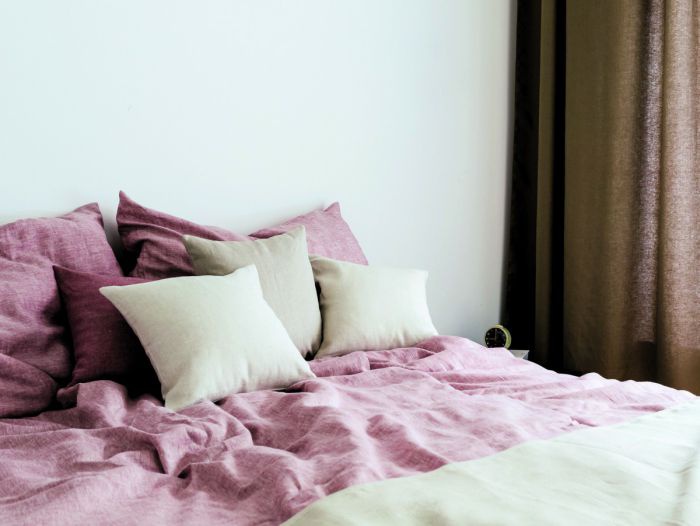 Ein helles Schlafzimmer, das Doppelbett neben dem großen Fenster ist mit Bettwäsche aus Leinen in Natur-, Bordeaux- und Rosétönen bedeckt.