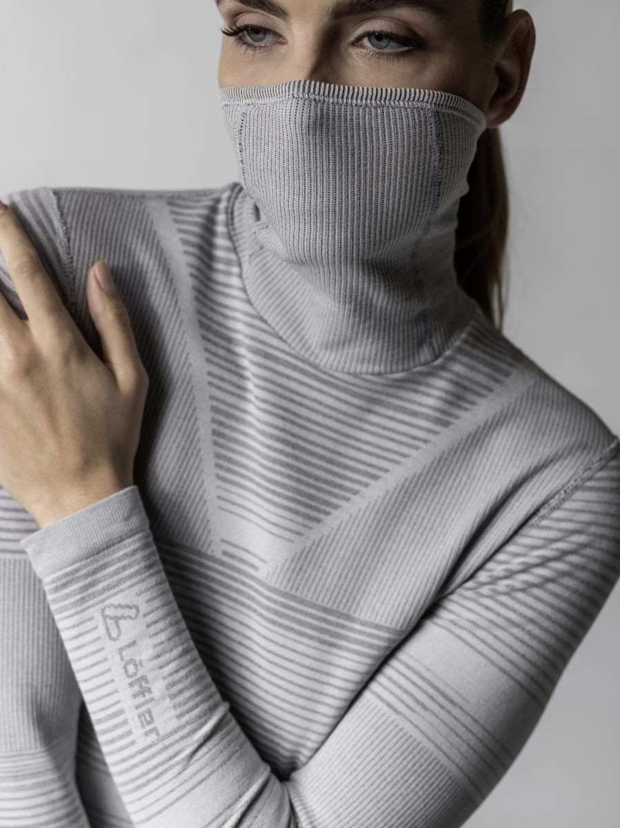Funktionsunterwäsche für den Winter: Ein weibliches Model trägt ein silbergraues, langärmeliges Oberteil mit Rollkragen, den sie bis über ihre Nase hoch aufgerollt hat und somit als Maske dient.