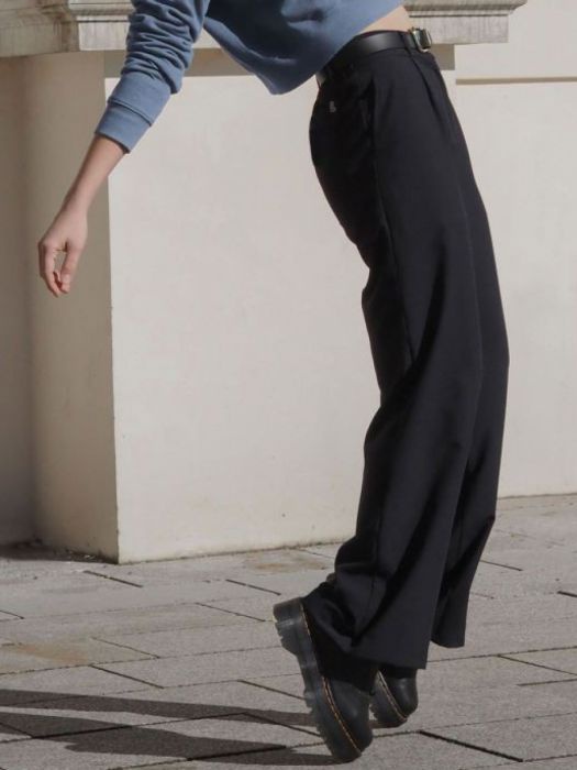 Ein weibliches Model mit Sonnenbrille posiert in taubenblauem Sweater und schwarzer Hose mit weitem Bein. Sie trägt dazu schwarze Stiefel mit Plateausohle und balanciert in nach hinten gebogener Körperhaltung auf den Zehenspitzen, die Arme seitlich nach o