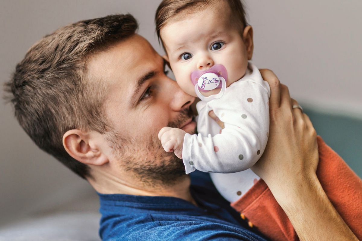 Ein junger Mann in blauem T-Shirt, mit kurzen braunen Haaren und kurzem Vollbart, hält ein Baby im Alter von einigen Monaten ganz nah an sich, sodass seine Nase die rechte Wange des Kindes berührt. Das Baby trägt zur orangefarbenen Strampelhose ein weißes