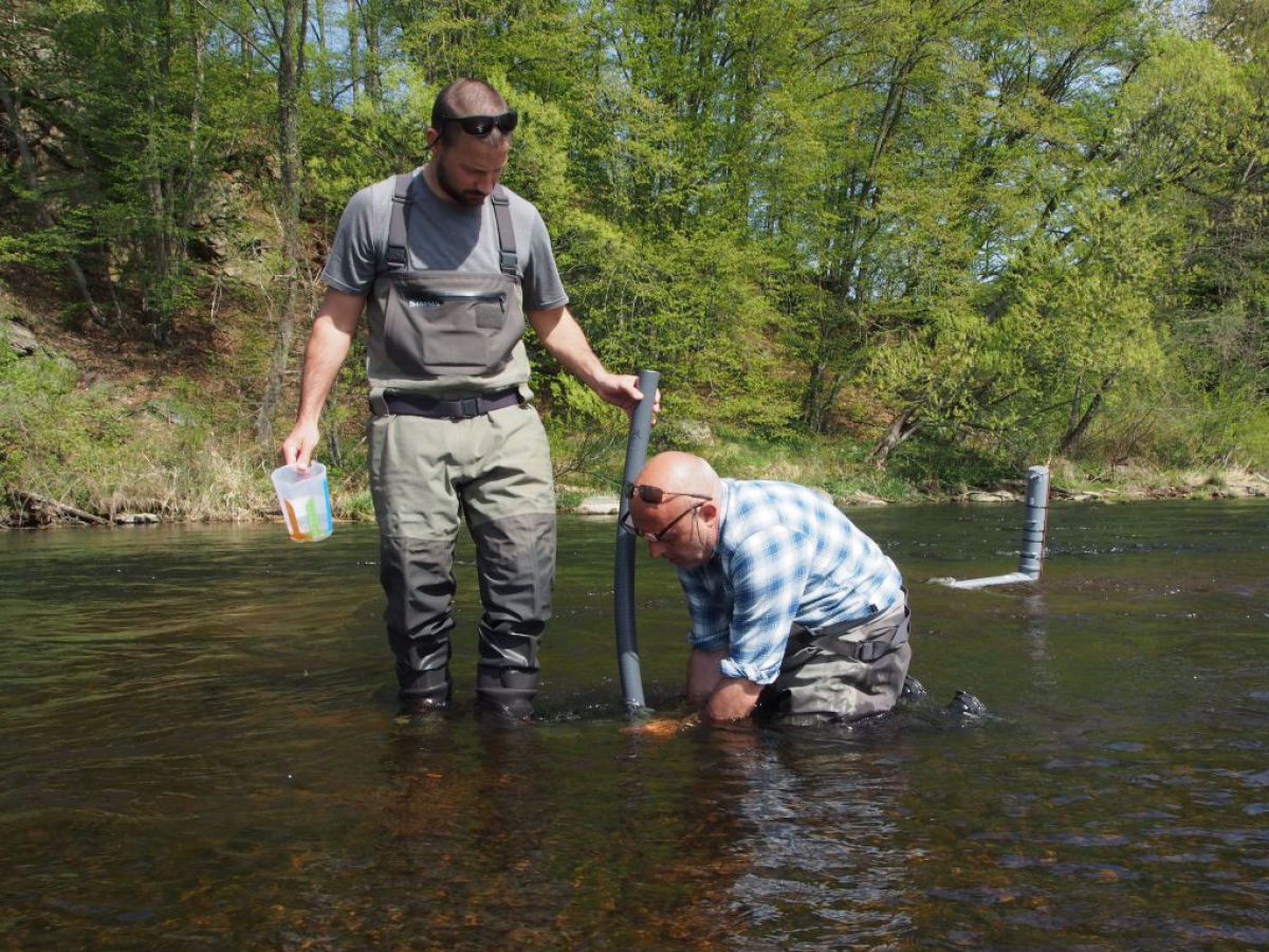 Zwei Männer in Watthosen hantieren im Fluss mit einem Rohr und anderen Behältern.