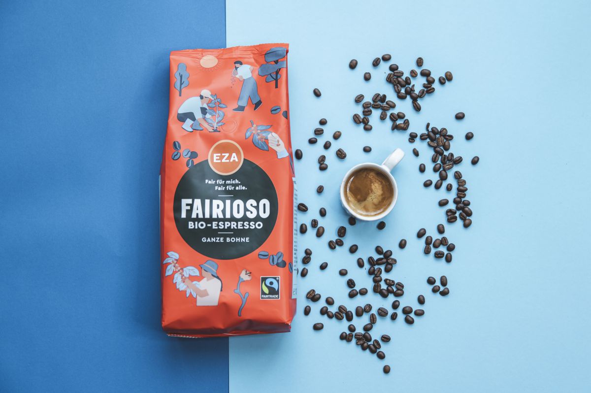 Eine rote Kaffeepackung mit bunten Zeichnungen  liegt vor blauem Hintergrund. Daneben eine Kaffeetasse und Kaffeebohnen.