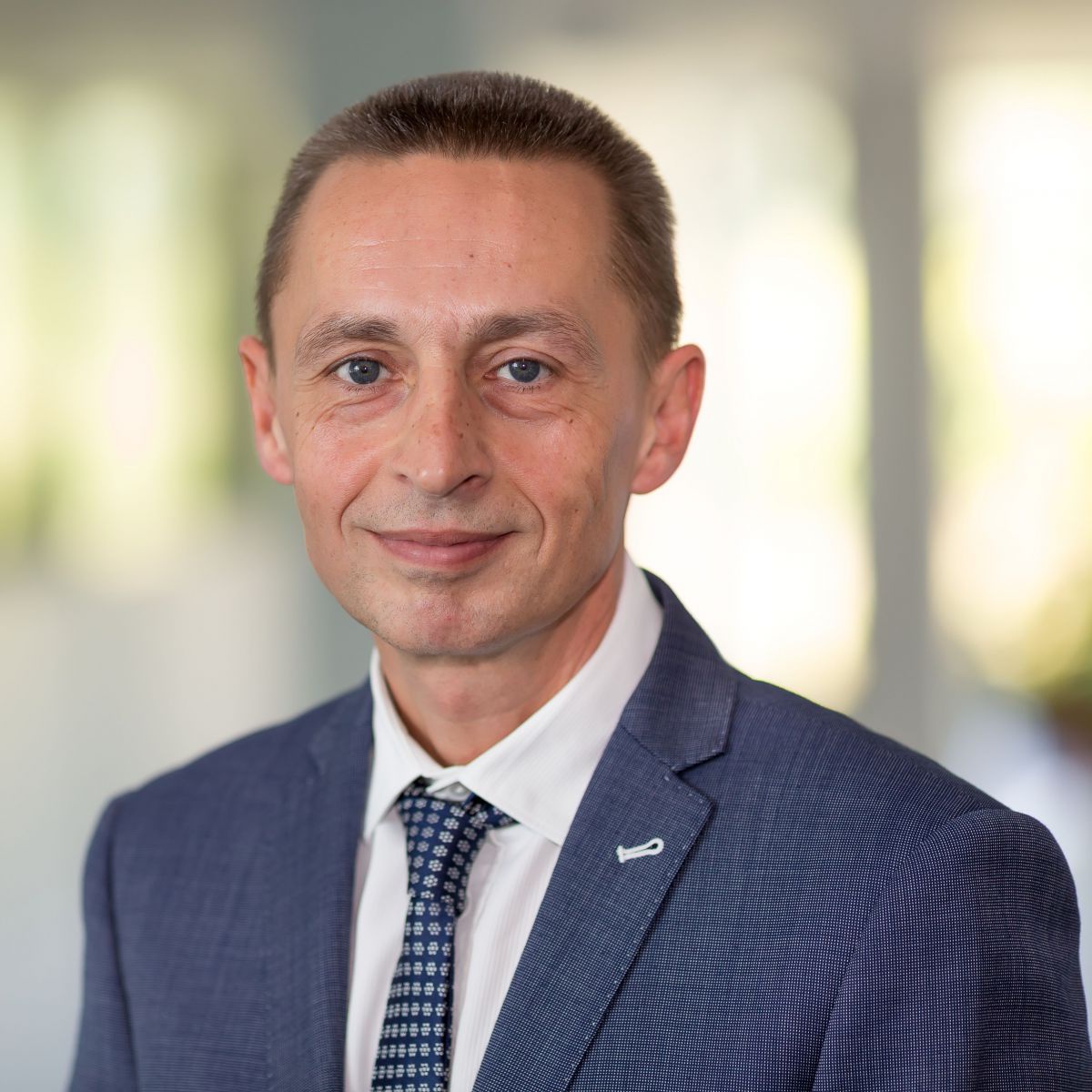 Ein Portraitfoto eines Mannes mit kurzen Haaren, blauen Augen, der einen Anzug trägt und in die Kamera lächelt.