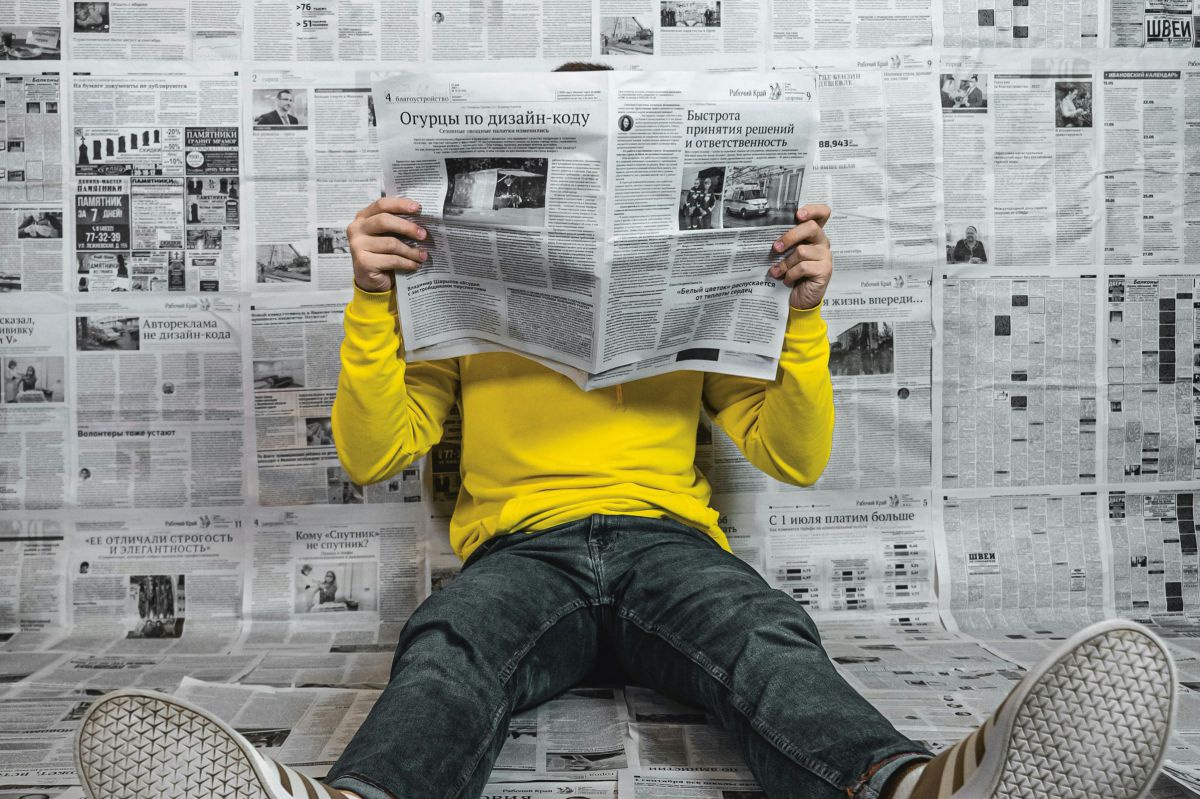 Ein Mann in leuchtend gelbem Pullover, schwarzen Jeans und Sneakers, sitzt mit ausgestreckten Beinen an eine Wand gelehnt, sein Gesicht ist hinter der Zeitung verborgen, in der er liest. Auch Boden und Wand sind gänzlich mit Zeitungsbögen tapeziert.