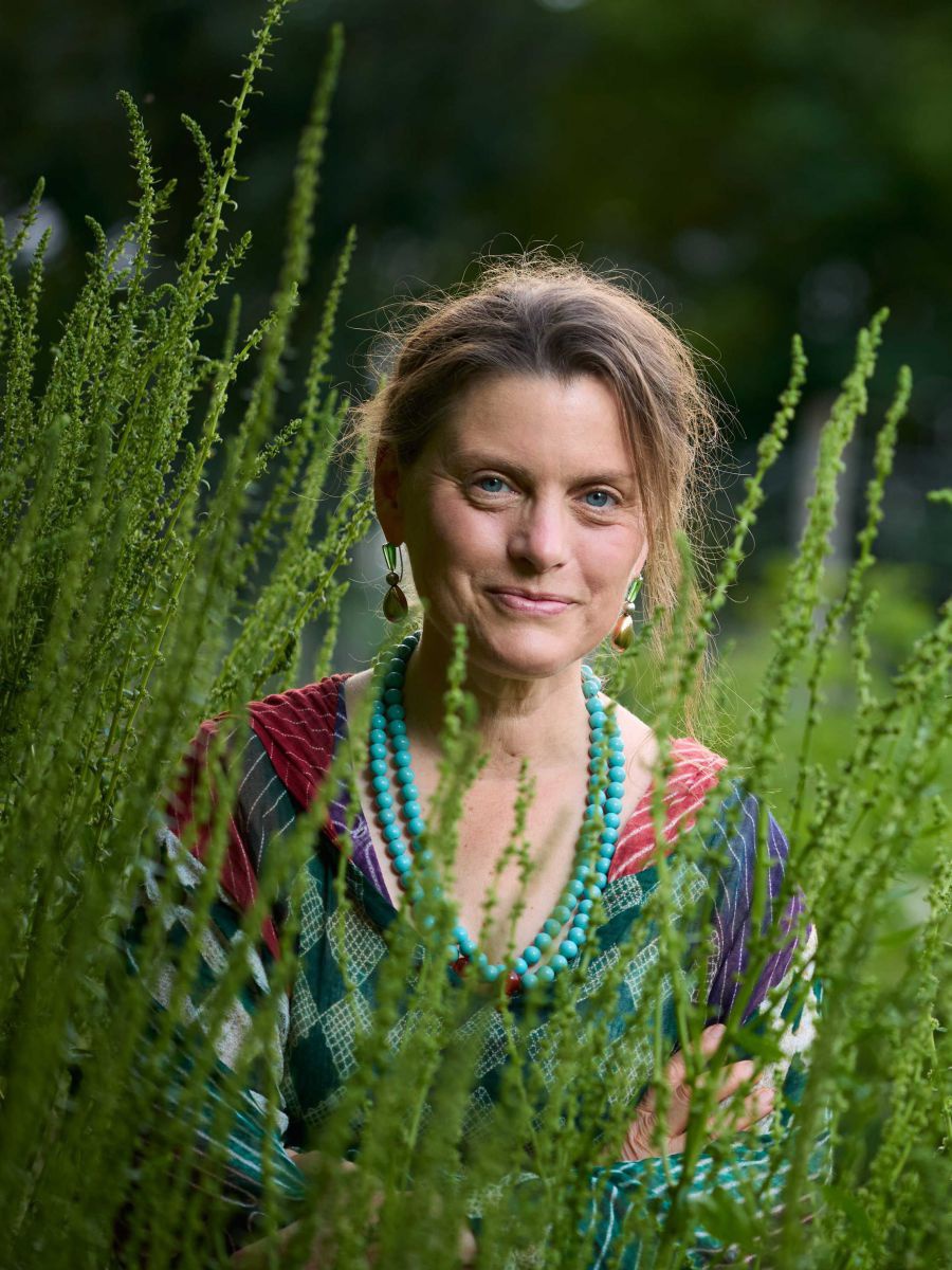 Gartenexpertin Andrea Heistinger sitzt in einer hochgewachsenen Wiese. Sie trägt ein bunt gemustertes Kleid und eine türkise, mehrreihige Steinperlenkette.
