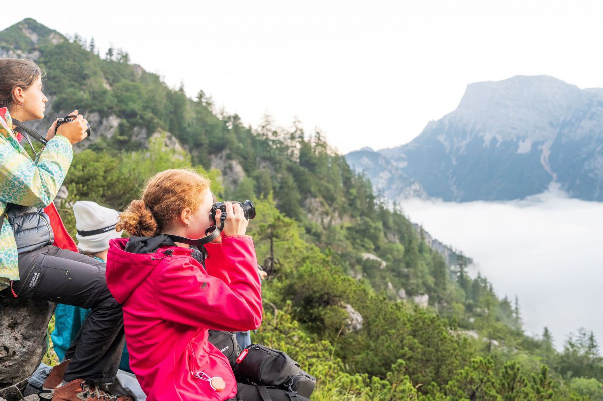 Zwei Jugendlige sitzen auf einem Hang in den Bergen und fotografieren in die Weite.