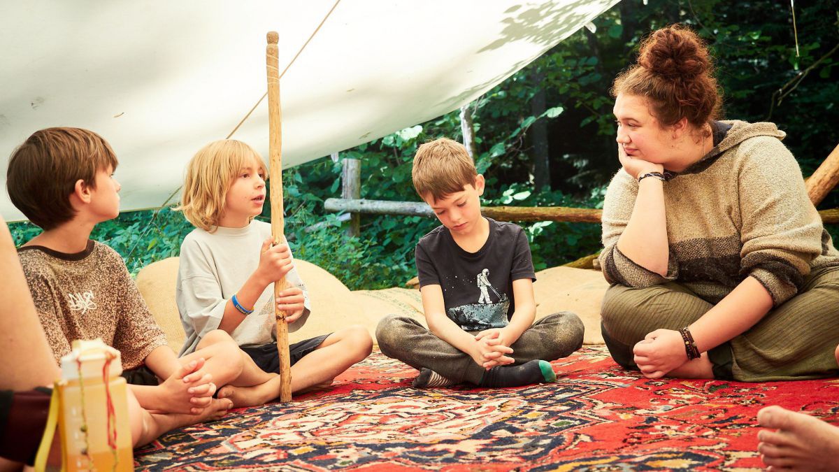 Kinder sitzen auf einem roten Teppich unter einer Sonnenplane im Freien. Eines davon hält einen Stock in Händen und spricht.