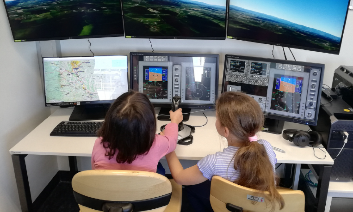 Zwei Mädchen sitzen vor Computerbildschirmen und steuern mit einem Joystick eine Flugmaschine.