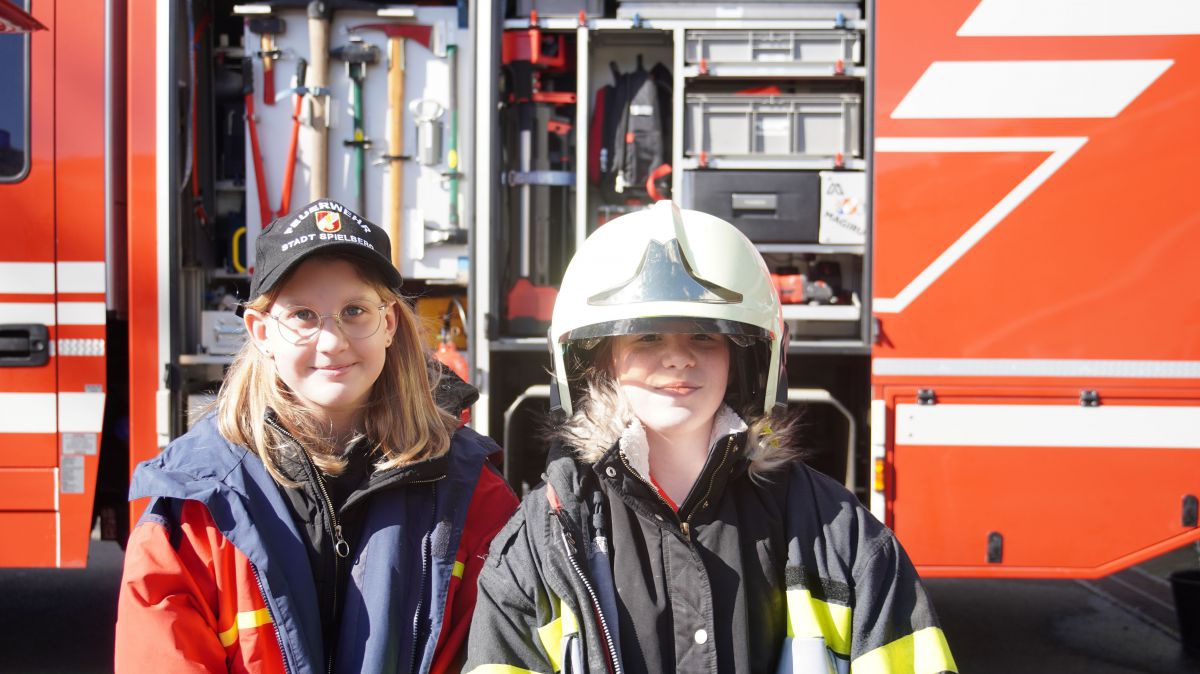 Zwei Mädchen stehen vor einem Feuerwehrauto. Sie tragen Feuerwehrjacken, eines hat einen Helm auf.