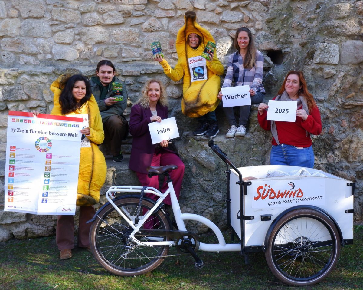Sechs Personen präsentieren die Fairen Wochen 2025. Sie sitzen auf bzw. stehen vor einer Steinmauer, zwei von Ihnen sind als fairtrade-Banane verkleidet, eine davon hält ein Plakat mit den 17 Zielen. Im Vordergrund steht ein weißes Lastenrad mit dem Südwi