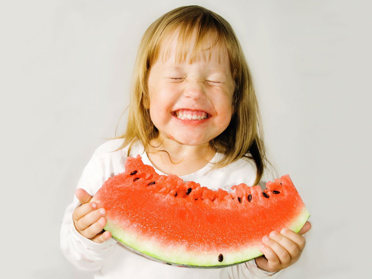 Ein Mädchen hat ein großes Stück Wassermelona in beiden Händen, von dem sie gerade einen Bissen genommen hat. Sie lacht mit vor Freude zusammengekniffenen Augen.