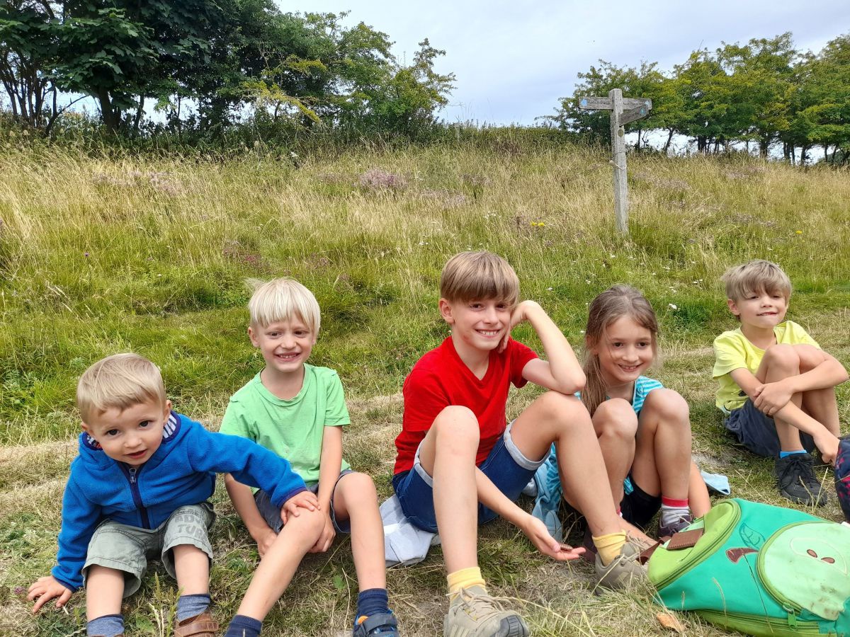 Fünf Kinder im Alter von etwa 2 bis 10 Jahren sitzen in sommerlicher Kleidung aufgereiht am Rand einer Wiese. Im Hintergrund ein paar Büsche.