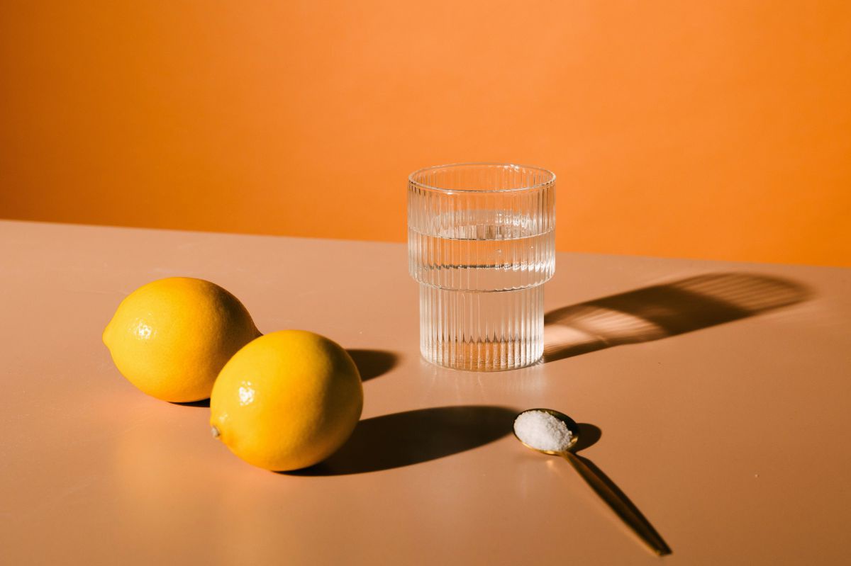 Vor einer orangenen Wand liegen zwei Zitronen auf einem beigen Tisch. Daneben ein Glas Wasser und ein Löffel mit weißem Pulver.