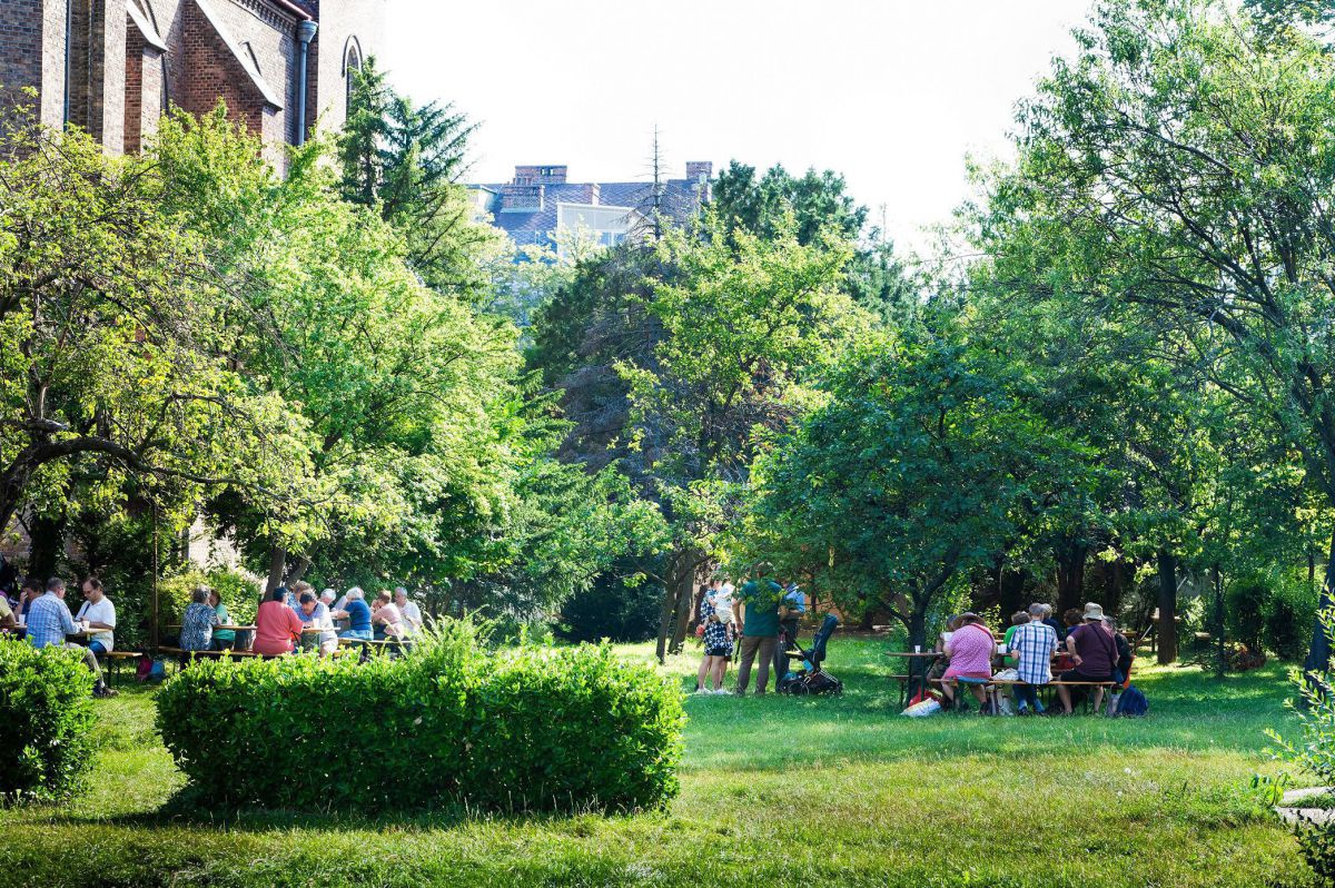 In einem städtischen Park, der an eine Kirche angrenzt, sitzen mehrere Gruppen von Menschen in Sommerkleidung an Sitzgelegenheiten im Schatten großer Bäume.