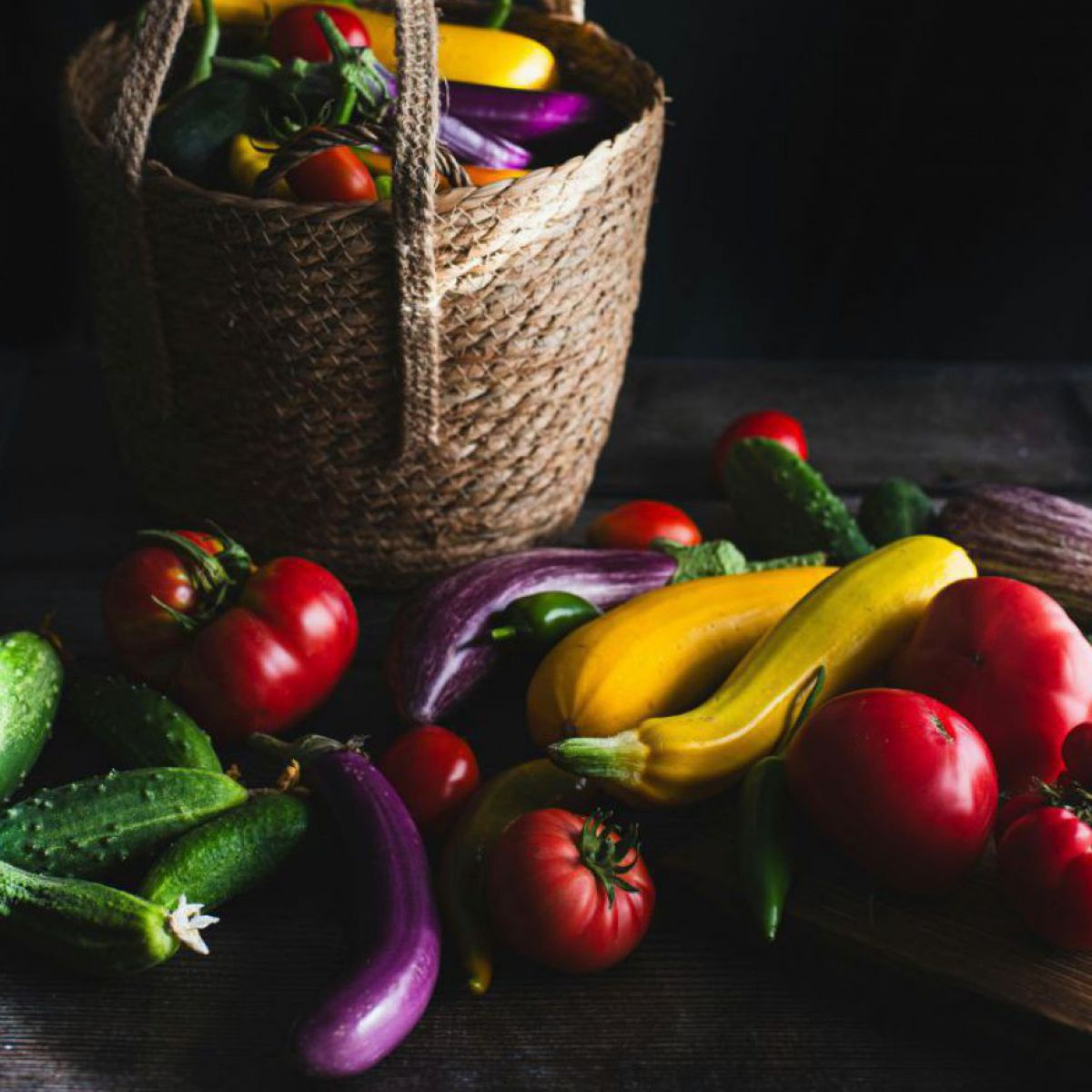 Ein Stilleben vor schwarzem Hintergrund: Eine Korbtasche aus Naturfaser, gefüllt mit Gemüse wie Tomaten, gelbe Zucchini, Feldgurken, Melanzani. Davor auf dem Tisch noch mehr dieser Gemüsesorten.