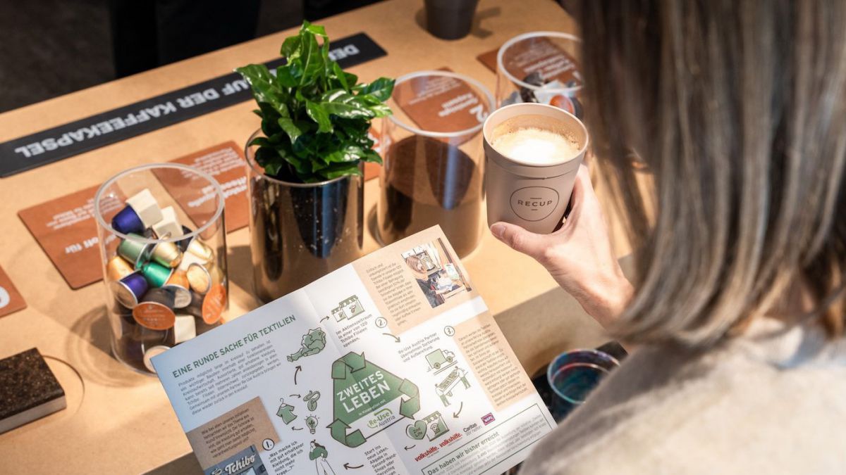 Eine Frau mit einem Kaffeebecher in der Hand liest eine Infobröschüre vor einem Display, das Kaffeekapselrecycling erklärt.