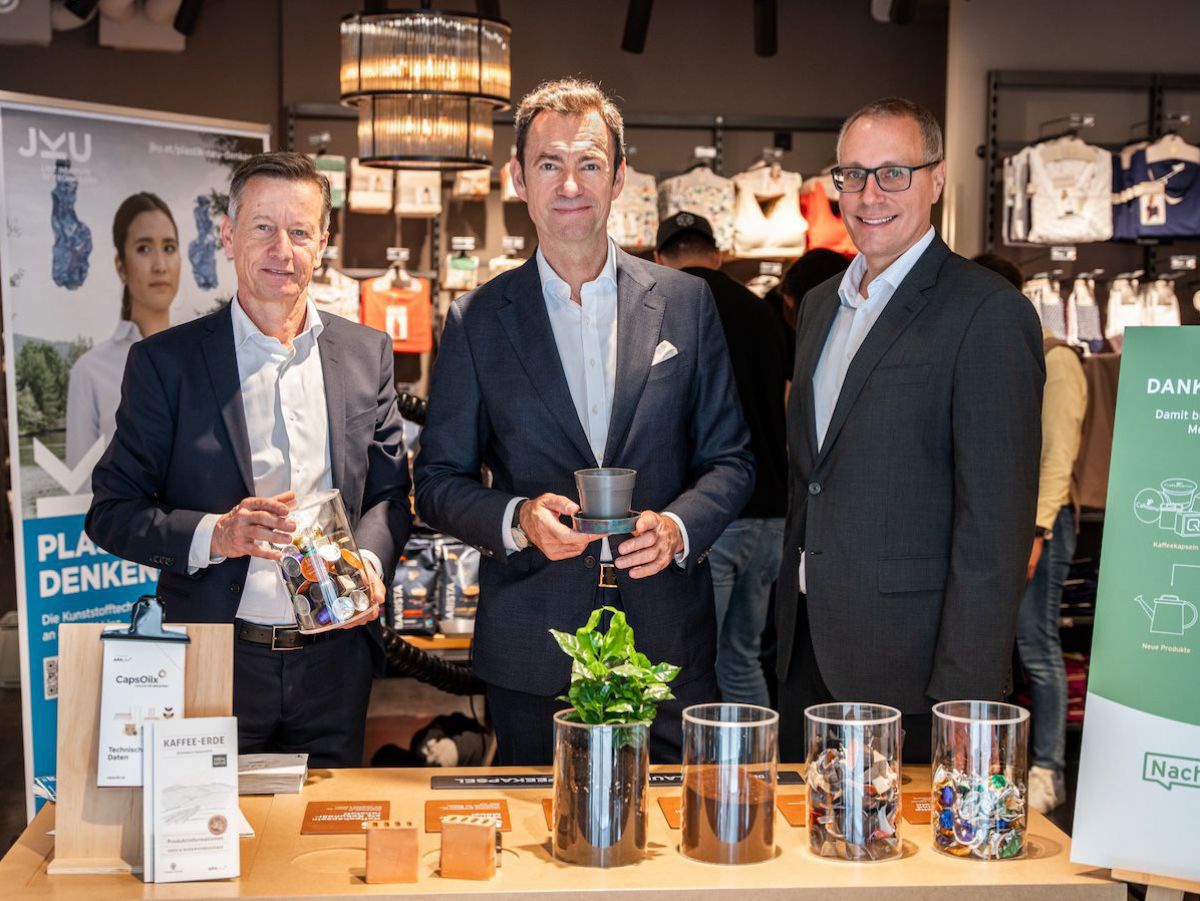 Drei Männer im Anzug stehen hinter einem Display, das Kaffeekapselrecycling erklärt.