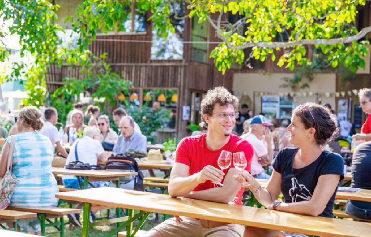 In einem Hof sind Heurigengarnituren aufgebaut, die meisten Tische sind mit Gästen belegt. Im Vordergrund sieht man ein Paar zu zweit an einem Tisch, sie stoßen mit Weißwein an. Über ihnen grün belaubte Zweige eines Baumes, durch die Blätter schimmert die