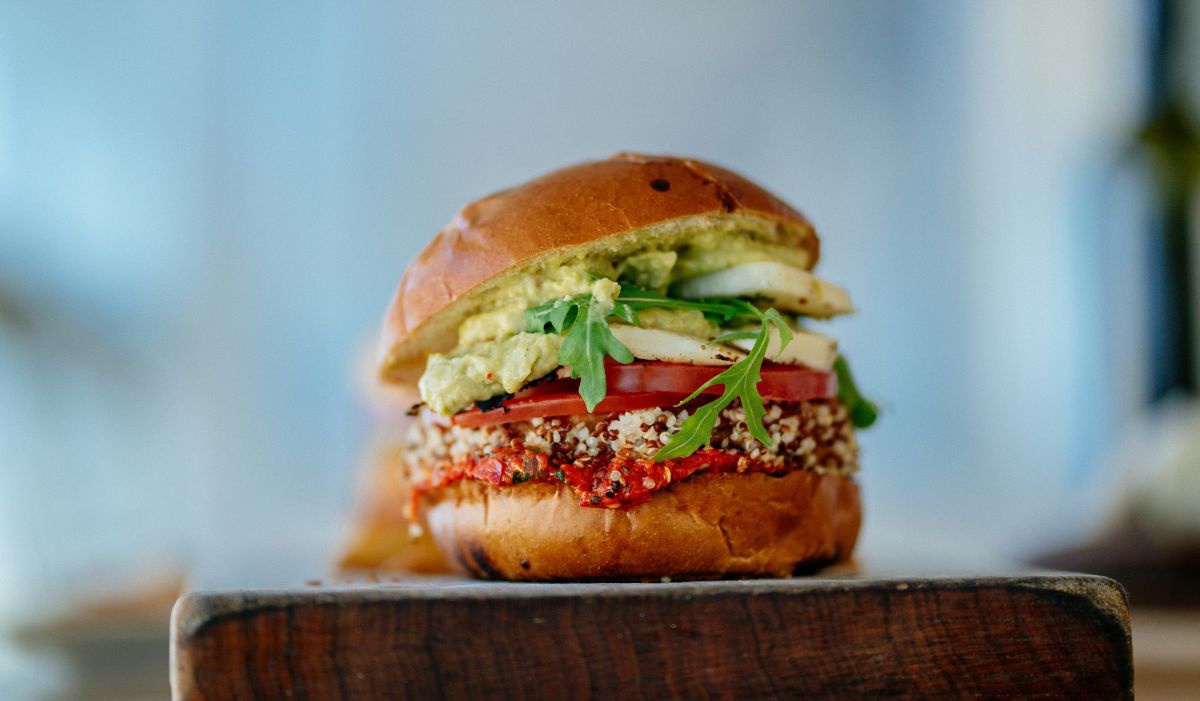 Ein veganer Burger mit Avocado und Tomate liegt auf einem Holzbrett. Er sieht appetitlich aus.