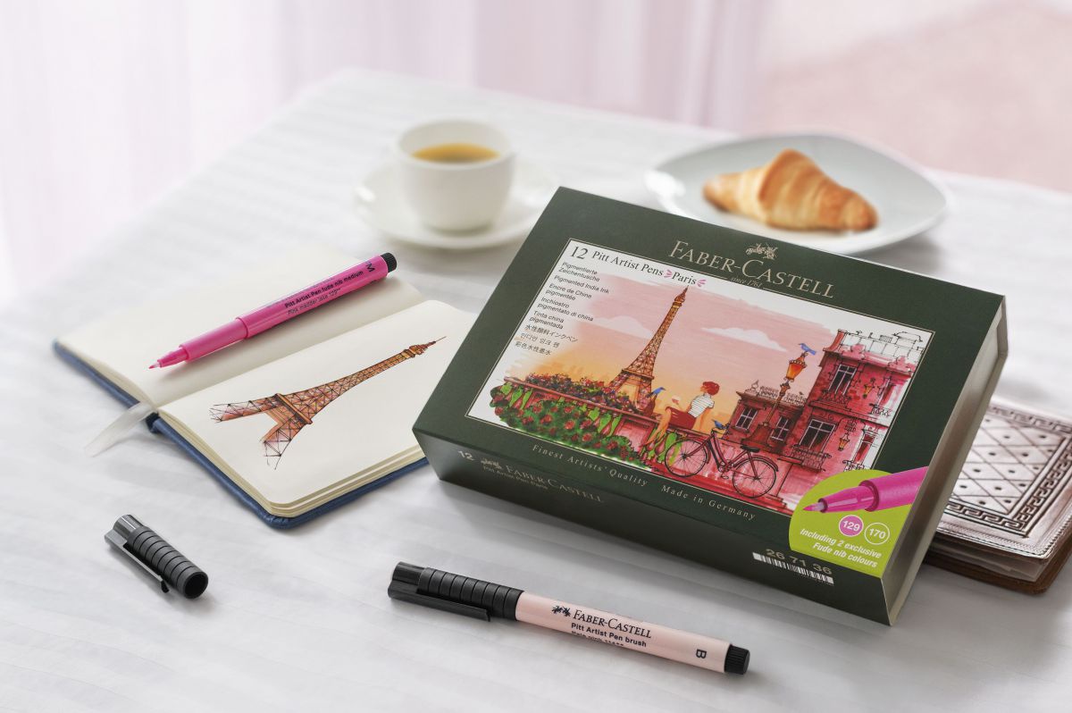 Auf einem Tisch steht ein Café und ein Croissant, davor ein Notizbuch, in dem der Eifelturm skizziert wird und Stifte sowie eine Stiftebox mit Paris-Motiv.