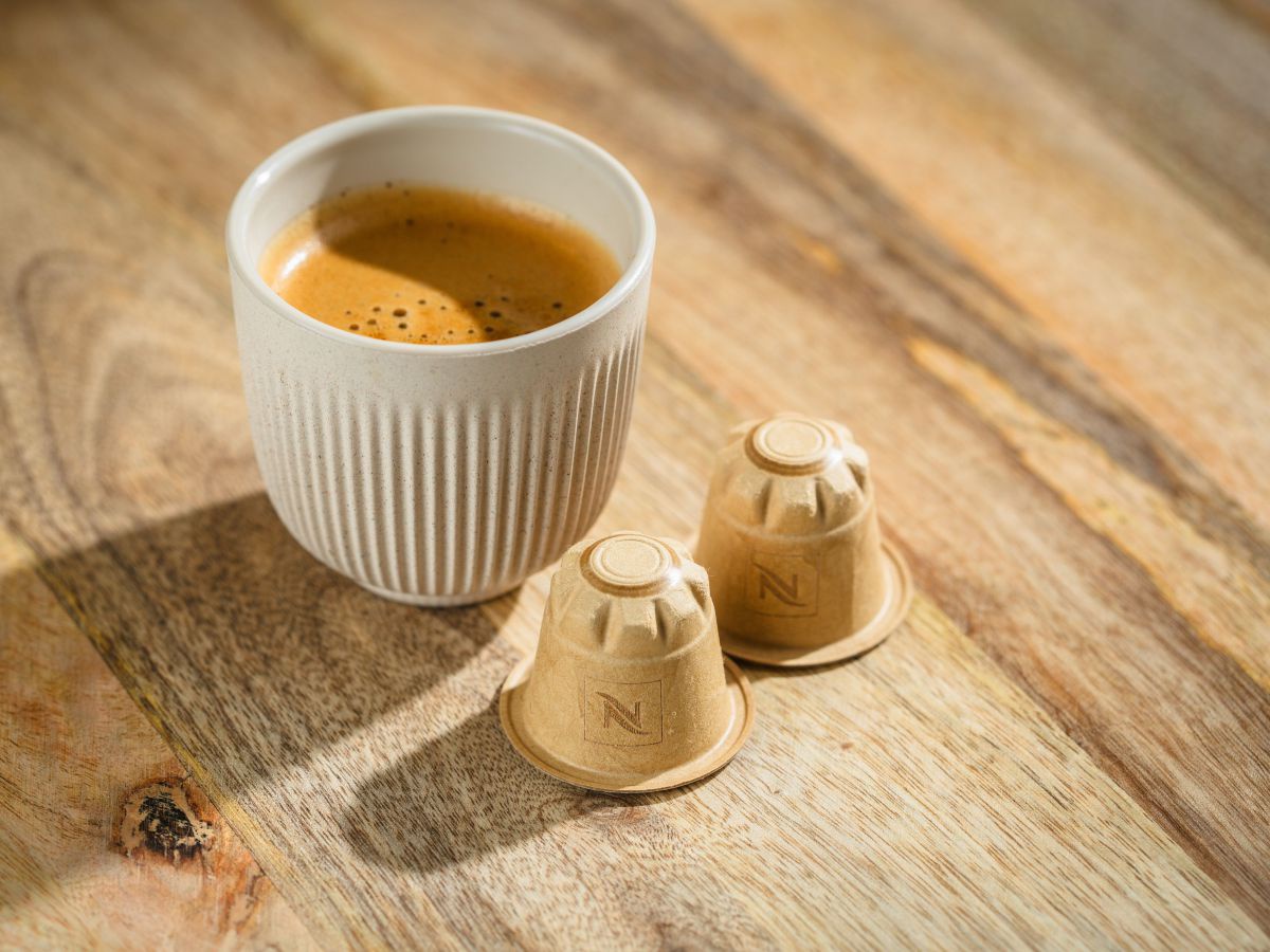 Auf einer Holztischplatte steht eine Tasse mit frisch gebrühtem Kaffee, daneben liegen zwei kompostierbare Kaffeekapseln.