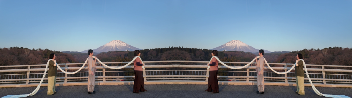 Ein Foto ab der Mitte gespiegelt zeigt drei Frauen, die ein Seil auf der Schulter liegen haben, und durch dieses verbunden sind. Im Hintergrund sieht man den Fuji.