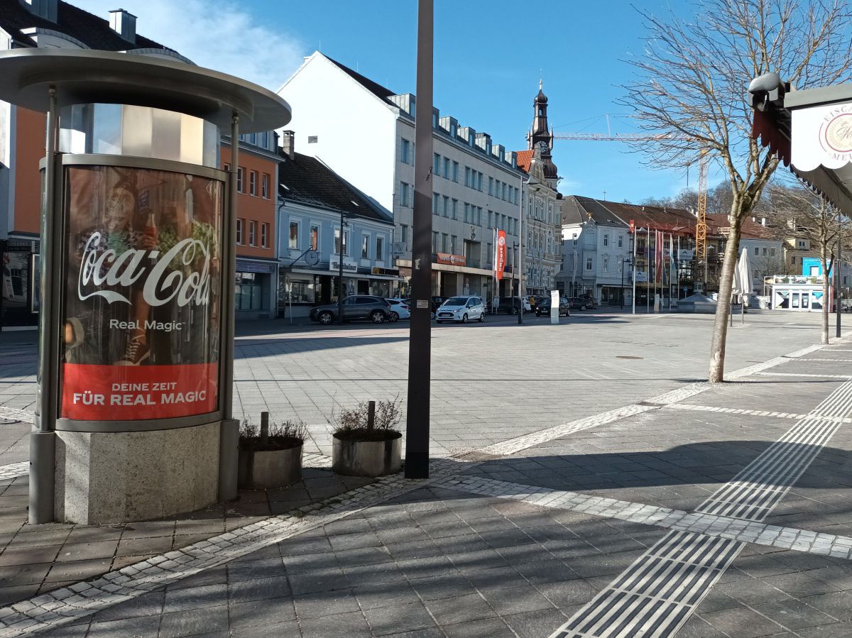 Blick auf einen Platz in der Stadt: außer ein paar schmächtigen Bäumchen gibt es hier nur Häuser und Autos auf Betonpflaster und Asphalt. Der Platz ist menschenleer. Links im Bild eine Litfasssäule mit Werbeplakat für eine weltbekannte Softdrinkmarke.