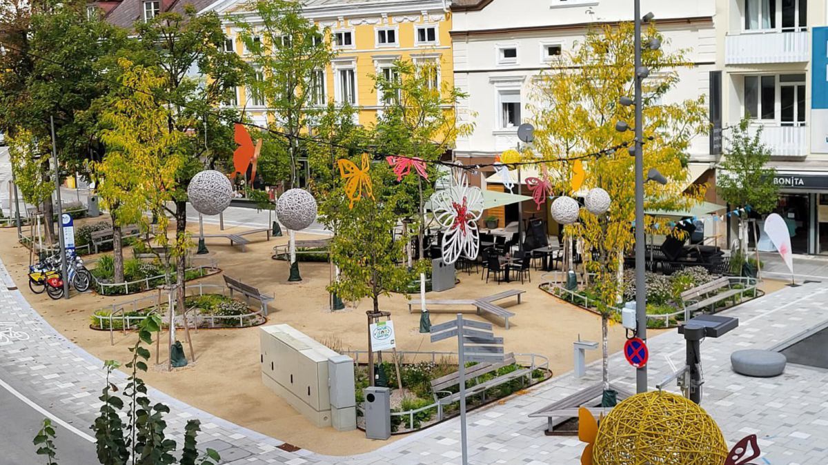 Blick auf einen Hauptplatz: zwischen Bäumen gibt es Blumenbeete und Wiesenelemente mit Sitzgelegenheiten und Fahrradständern. Über allem hängen Lampions und andere sommerliche Dekoration. Auch der Schanigarten eines an