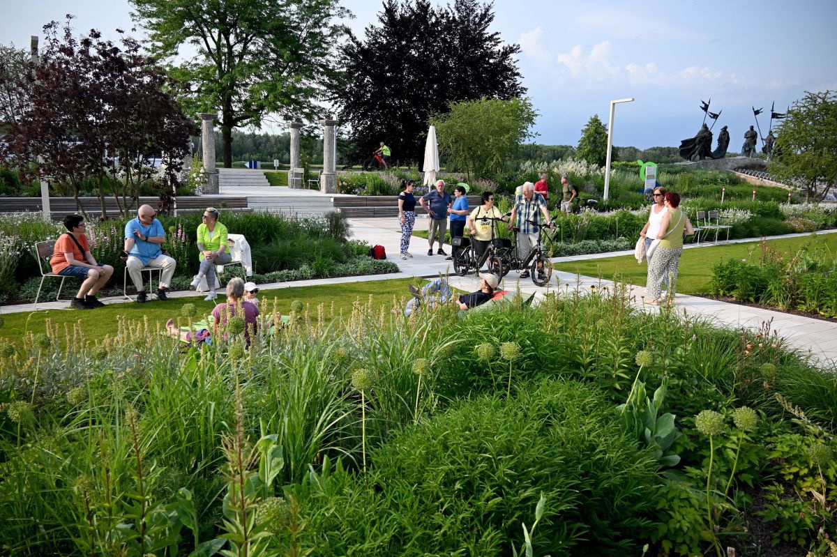 Blick in einen Park mit mehreren Ebenen und grünen Flächen mit verschiedenstem Bewuchs. Zwischen Rasen, Blumenbeeten, Büschen und Bäumen spazieren Menschen, fahren Rad oder sitzen auf Bänken oder in der Wiese.