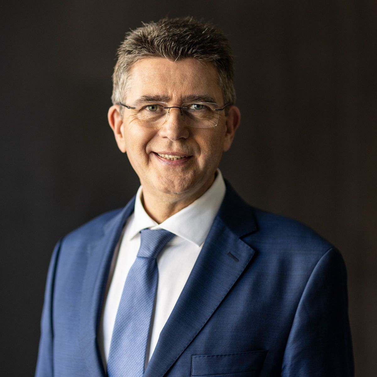 Ein Mann mittleren Alters im blauen Anzug mit weißem Hemd und blauer Krawatte. Er trägt die an den Schläfen angegrauten Haare kurz geschnitten und eine Brille.