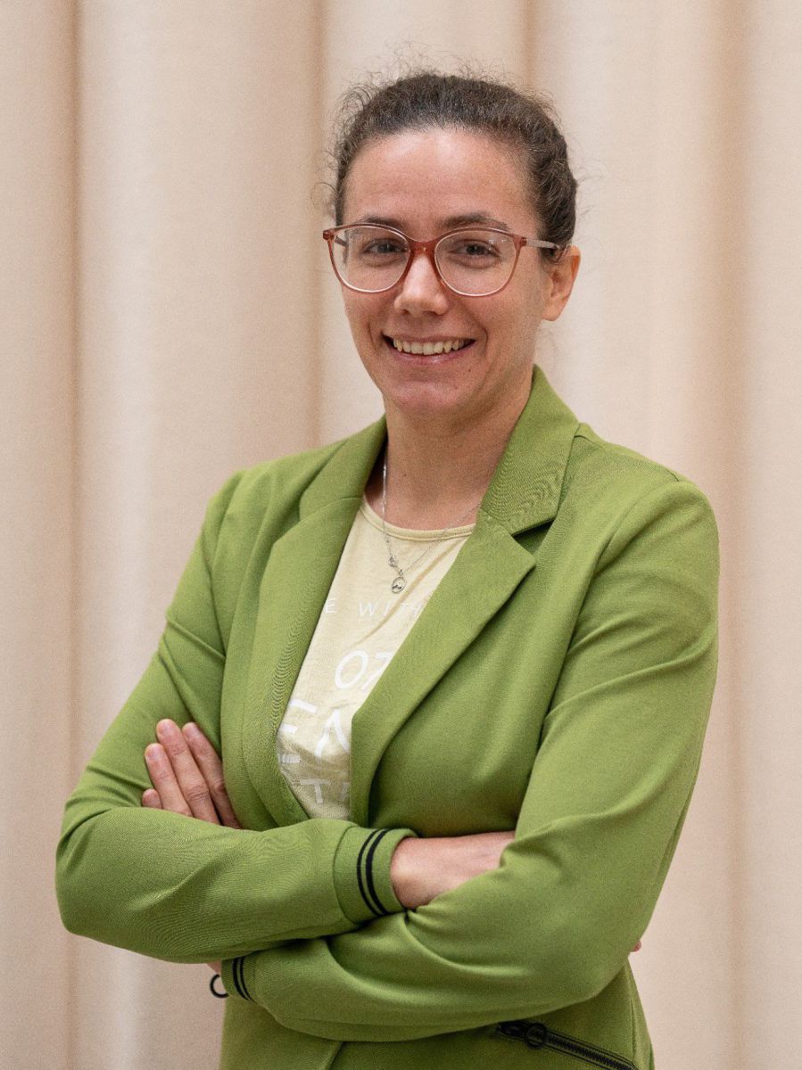 Eine Frau in etwa ihren Dreißigern steht mit verschränkten Armen vor einem hellen Vorhang und lächelt. Sie trägt einen olivgrünen Blazer über einem beigen Shirt, ihr krauses Haar hat sie nach hinten gebunden und ihre Brille hat einen bernsteinfarbenen Kun