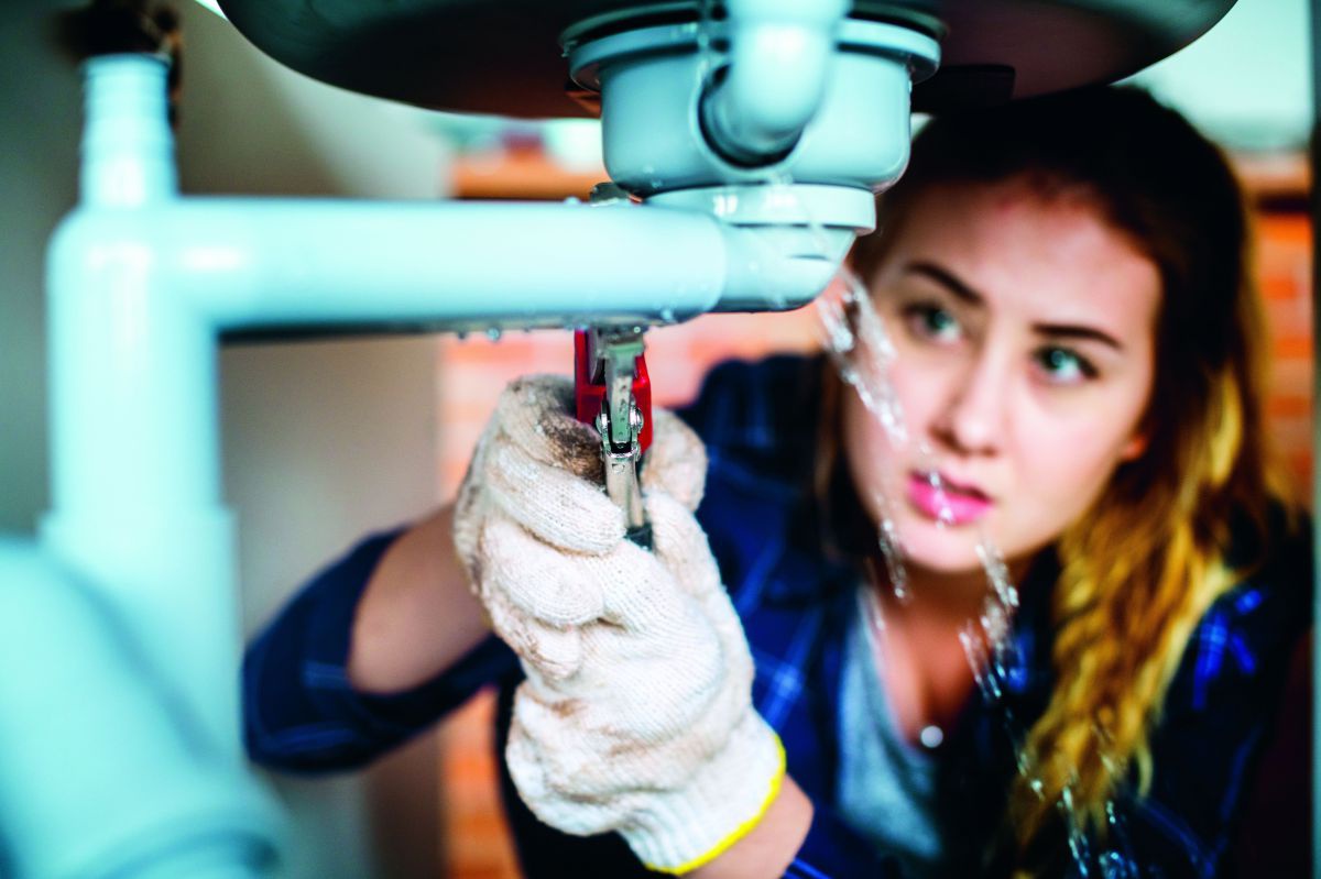Eine junge Frau hantiert mit einer Rohrzange am Abfluss unter einem Waschbecken. Wasser spritzt aus den Rohrverbindungen.