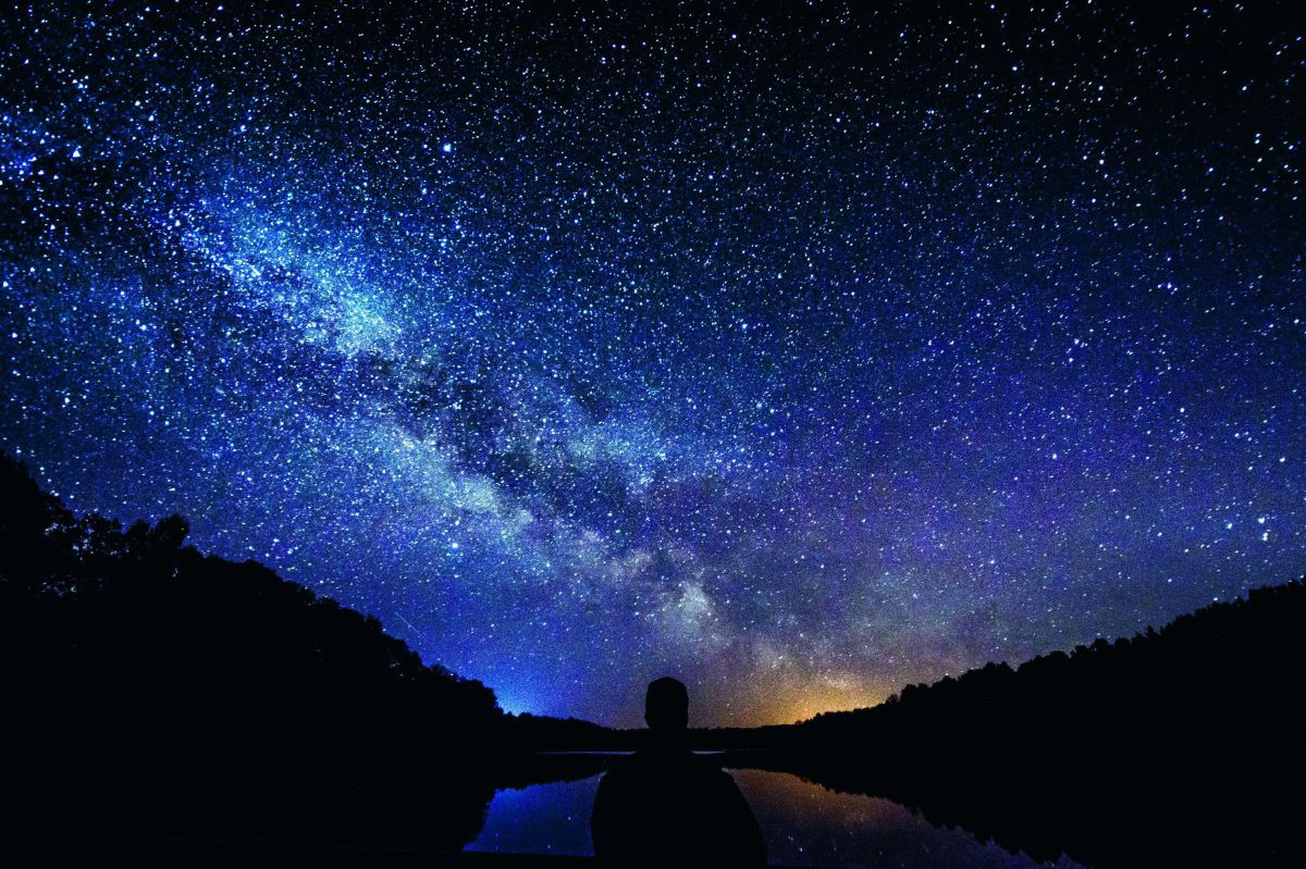Ein prachtvoller Nachthimmel, übersäht mit Sternen. Im Vordergrund sieht man die Silhouette einer Person vor einem See, in dessen Wasserfläche sich der Himmel spiegelt.