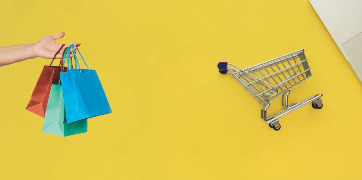 Ein senfgelber Hintergrund auf dem ein kleiner Einkaufswagen liegt und von der Seite bunte Papiertaschen ins Bild gehalten werden.