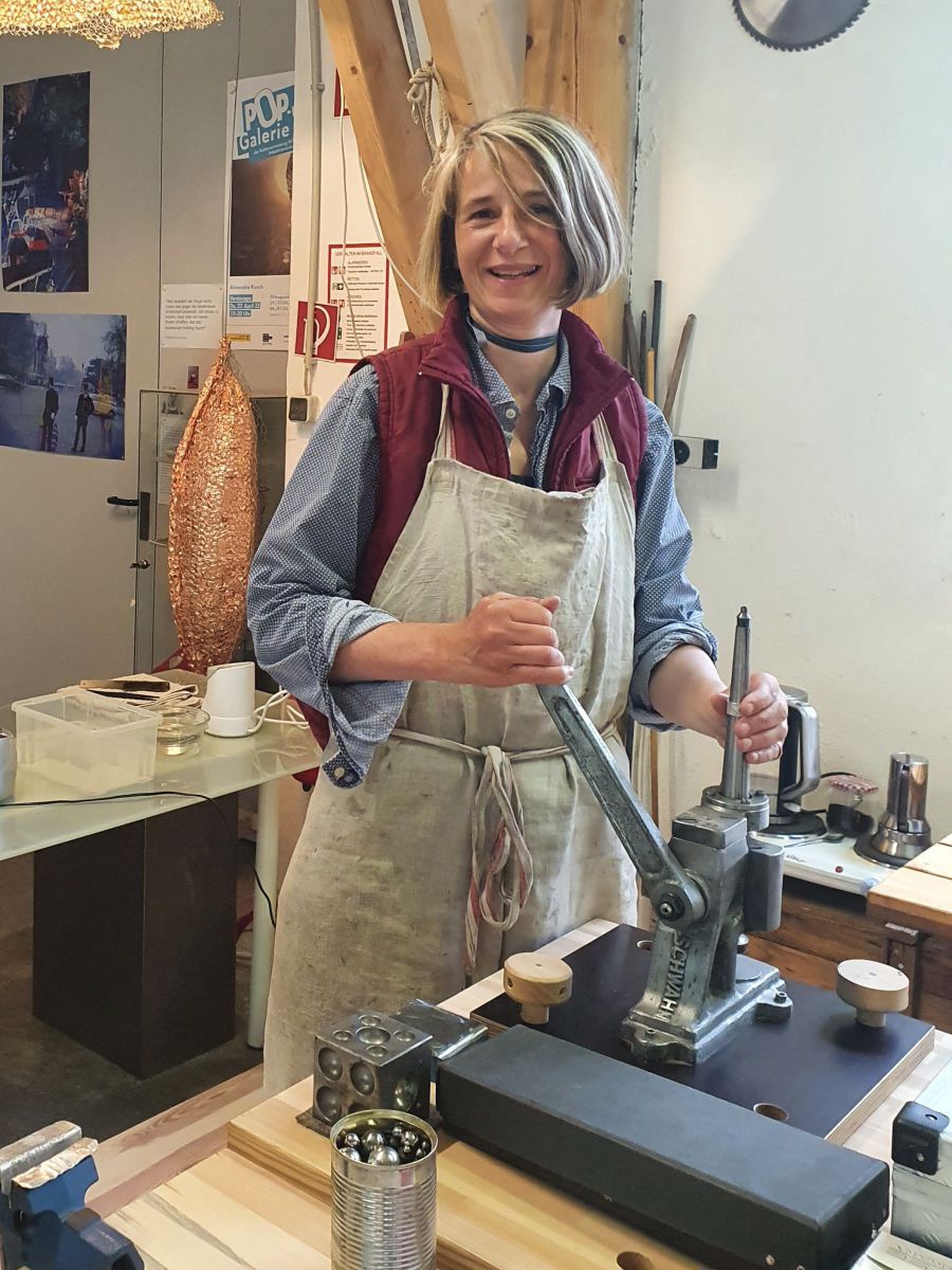 Eine Frau mit kinnlangen Haaren und einer Arbeitsschürze steht an einer Art Presse in einer Werkstatt.