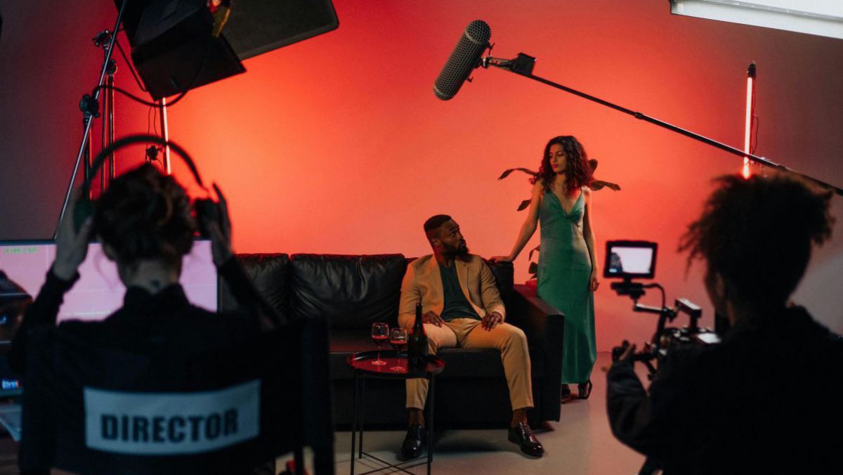 Das Bild zeigt einen Dreh in einem Studio. Das Set ist orangerot beleuchtet, ein Mann sitzt auf einem schwarzen Ledersofa, daneben steht eine Frau in grünem Kleid. Links im Vordergrund eine Frau mit Kopfhörern vor einem Laptop, auf der Stuhllehne steht 