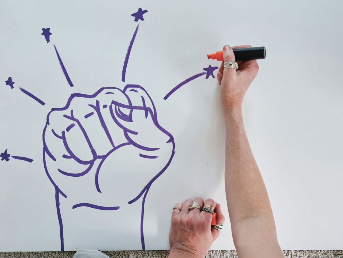 Eine Person, von der man nur die Hände, Arme und die Zehenspitzen in Socken sieht, sitzt auf einem hellen, flauschigen Teppichboden und gestaltet Transparente für eine Demo. An den Händen hat die Person Ringe an fast jedem Finger, in der rechten Hand hält