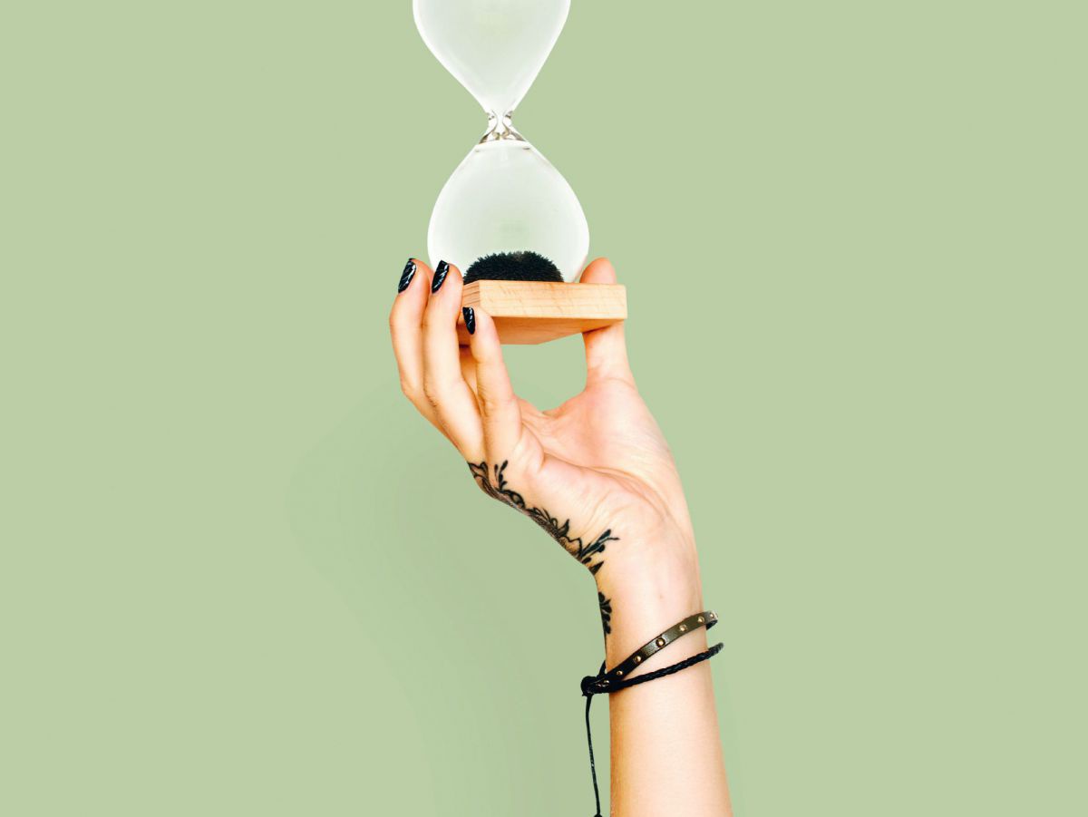 Eine Person, von der man nur rechte Hand und den Unterarm sieht, hält eine Sanduhr mit hellem Holzsockel ins Bild. Der Sand im Glas ist schwarz, ebenso wie die lackierten Fingernägel, das Tatoo am Handrücken und die Armbänder der Person. Der Hintergrund i