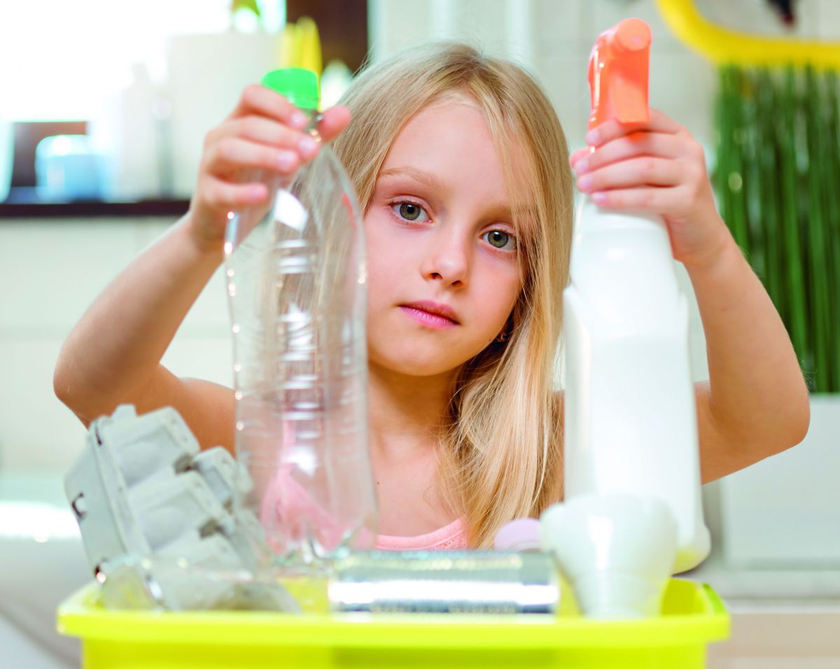 Ein Mädchen im Volksschulalter mit blonden langen Haaren steht in einem Badezimmer und hält in der linken Hand eine leere PET-Flasche und in der rechten eine Sprühflasche ohne Etikett.