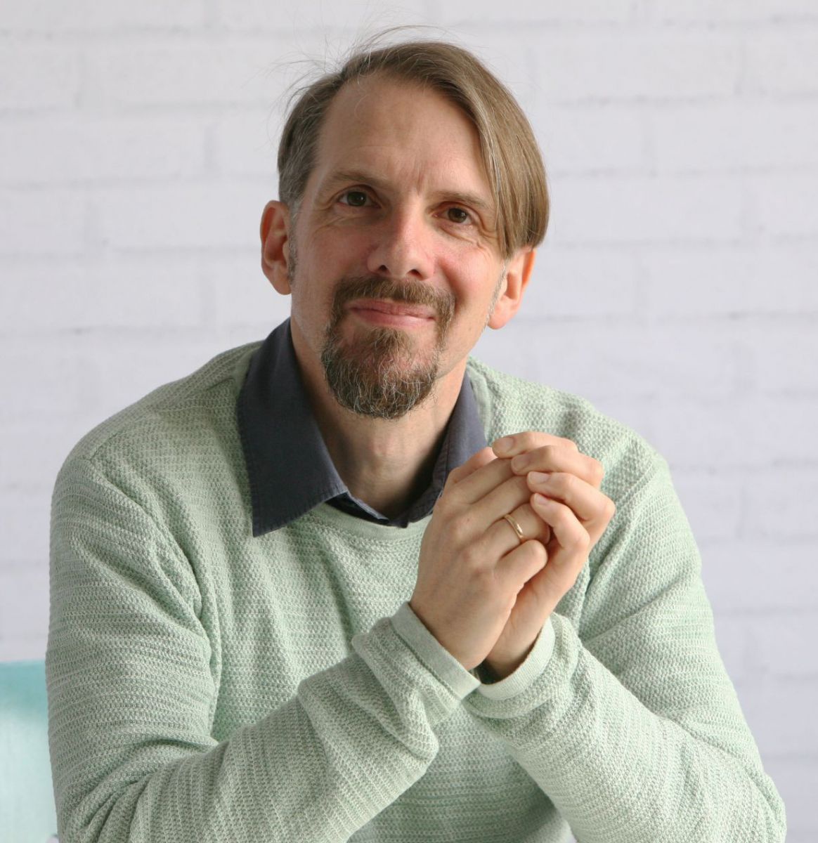 Portraitfoto: Mann um die Vierzig, glattes braunes Haar bis zu den Ohren, seitlich über den Kopf frisiert, Henri-Quatre-Bart, sitzt an einem Tisch, Ellbogen aufgestützt und Hände verschränkt. Er trägt einen hellen Pullover, der Kragen eines grauen Hemdes 