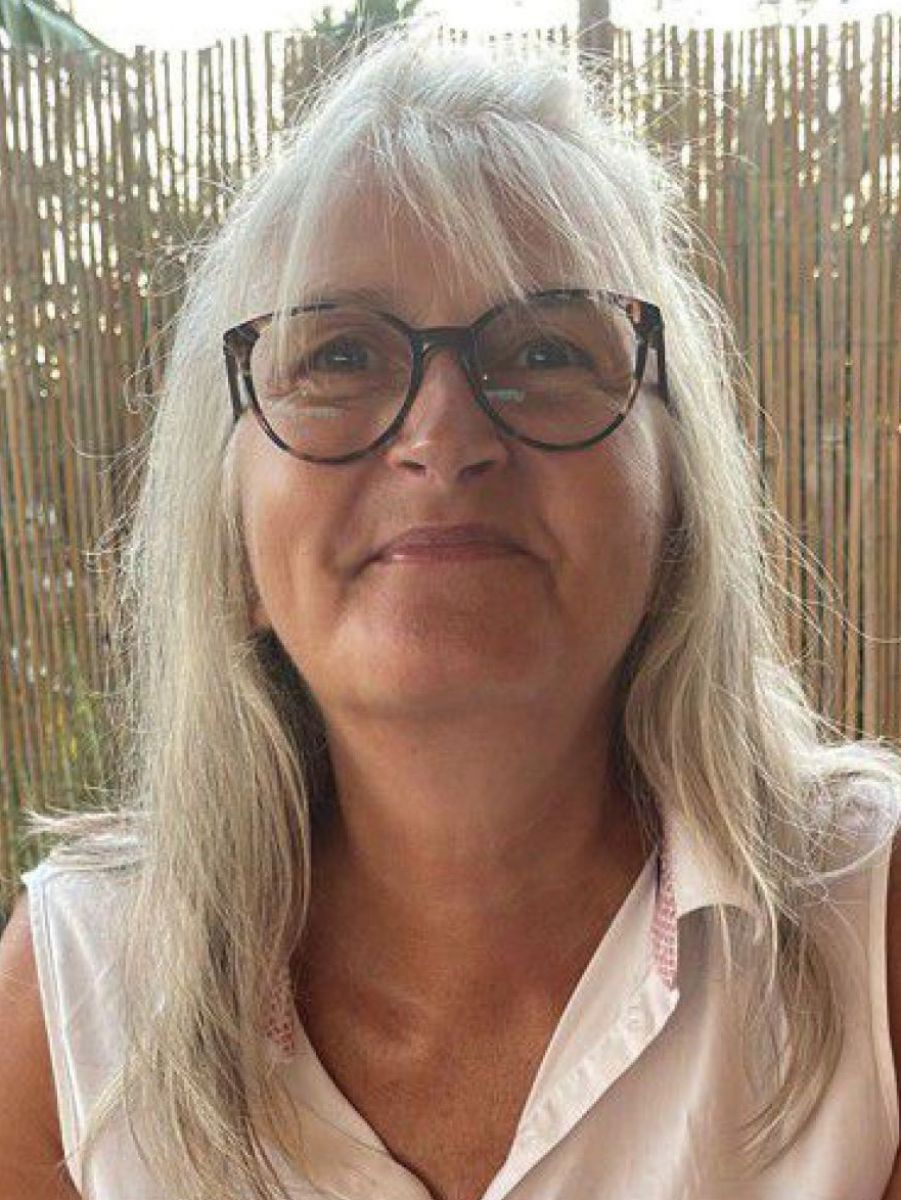 Portraitfoto: Frau in ihren Fünfzigern lächelt in die Kamera, langes graublondes Haar mit Stirnfransen, große Brille, ärmellose weiße Bluse. Schilfmatte im Hintergrund.