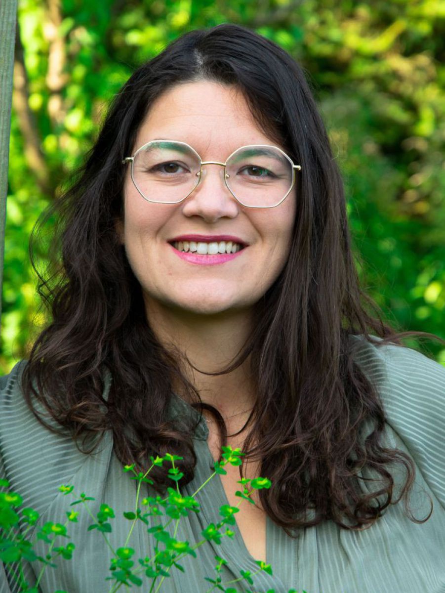 Portraitfoto: Frau in ihren Dreißigern, offenes, schwarzbraunes, langes Haar, dunkle Augen, Brille mit achteckigem, goldenen Rahmen, offenes Lächeln, trägt graugrüne Faltenbluse. Im Hinter- und Vordergrund Sträucher und Pflanzen.