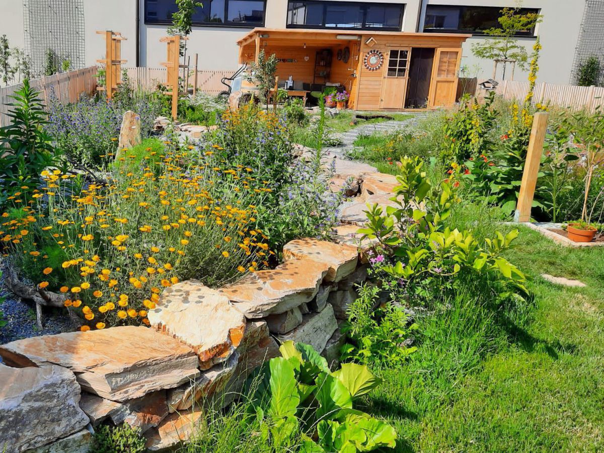 Halbhohe Mauer aus aufgeschichteten Steinen als Begrenzung für ein Hochbeet. Die Pflanzen blühen, Kürbispflanzen ranken sich hinunter bis auf die Wiese. Hölzernes Gartenhaus und weitere üppig bewachsene Beete im Hintergrund.