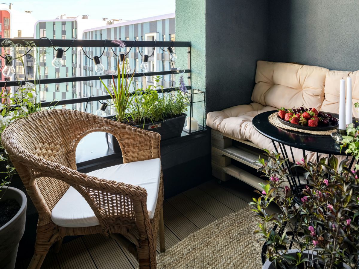 Blick auf einen gemütlch eingerichteten Balkon mit Pflanzen in Töpfen und Trögen, Rattansessel, Palettensofa, Sisalteppich, rundes Tischchen mit Kerzen und Teller mit Erdbeeren darauf.