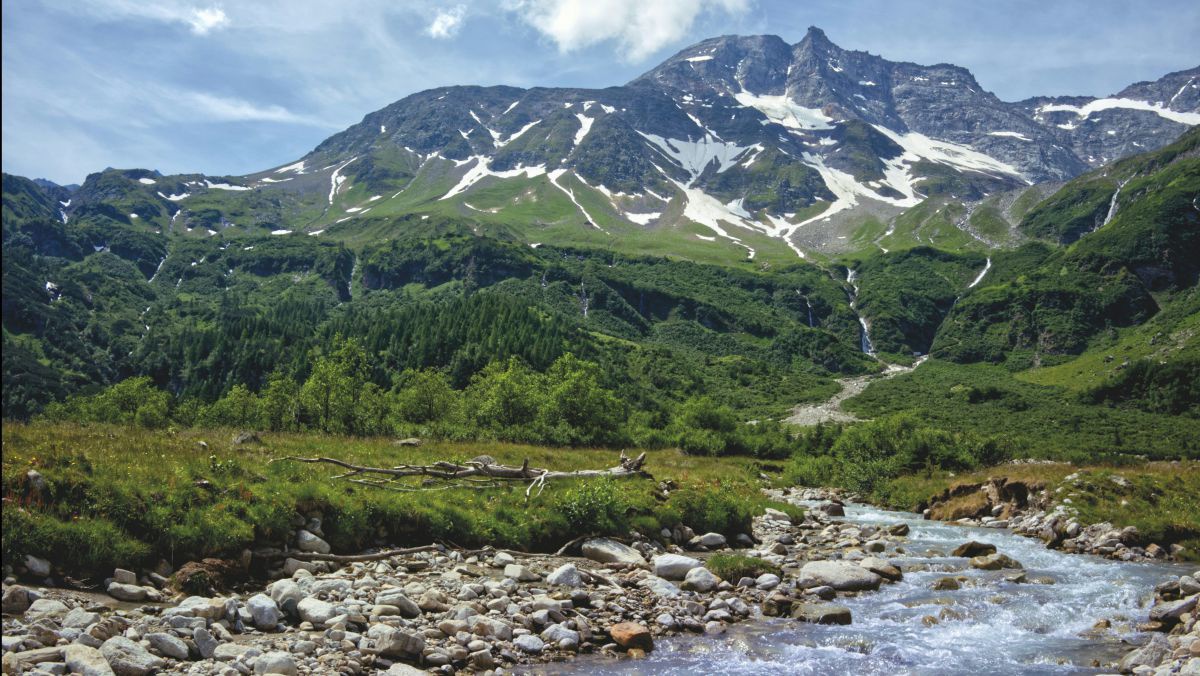 Ein Bach plätzschert im Vordergrund, dahinter grüne Landschaft und ein Berg auf dem sich noch letzte Schneereste finden.