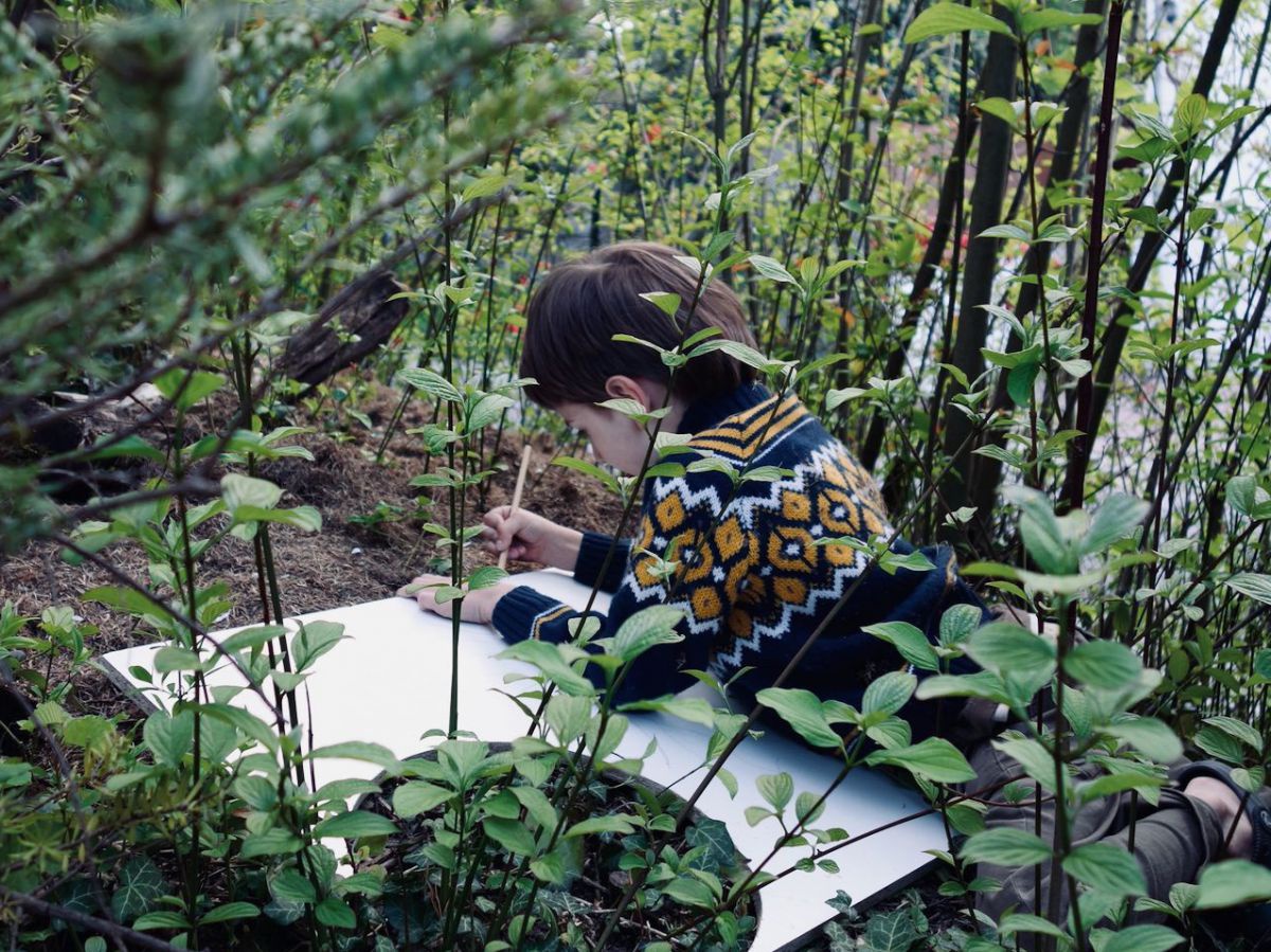 Ein Kind liegt mit einem großen Block zwischen Pflanzen im Garten und zeichnet.
