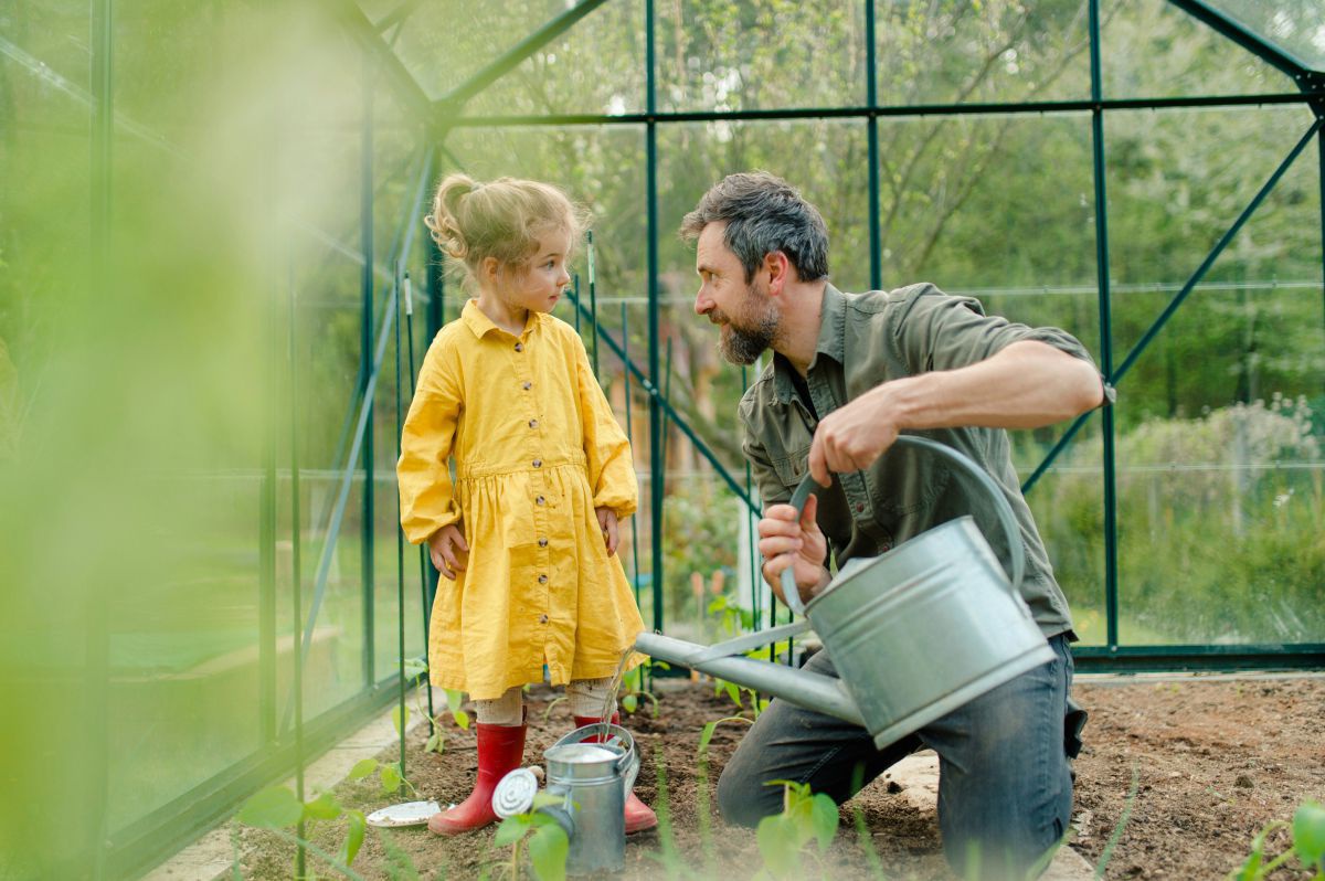 Ein Kind mit gelben Kleid und roten Stiefeln steht in einem Glashaus neben einem Mann der kniet und mit seiner großen Gießkanne eine kleinere für das Kind anfüllt.